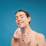A smiling woman applying lip gloss with closed eyes on a blue background.
